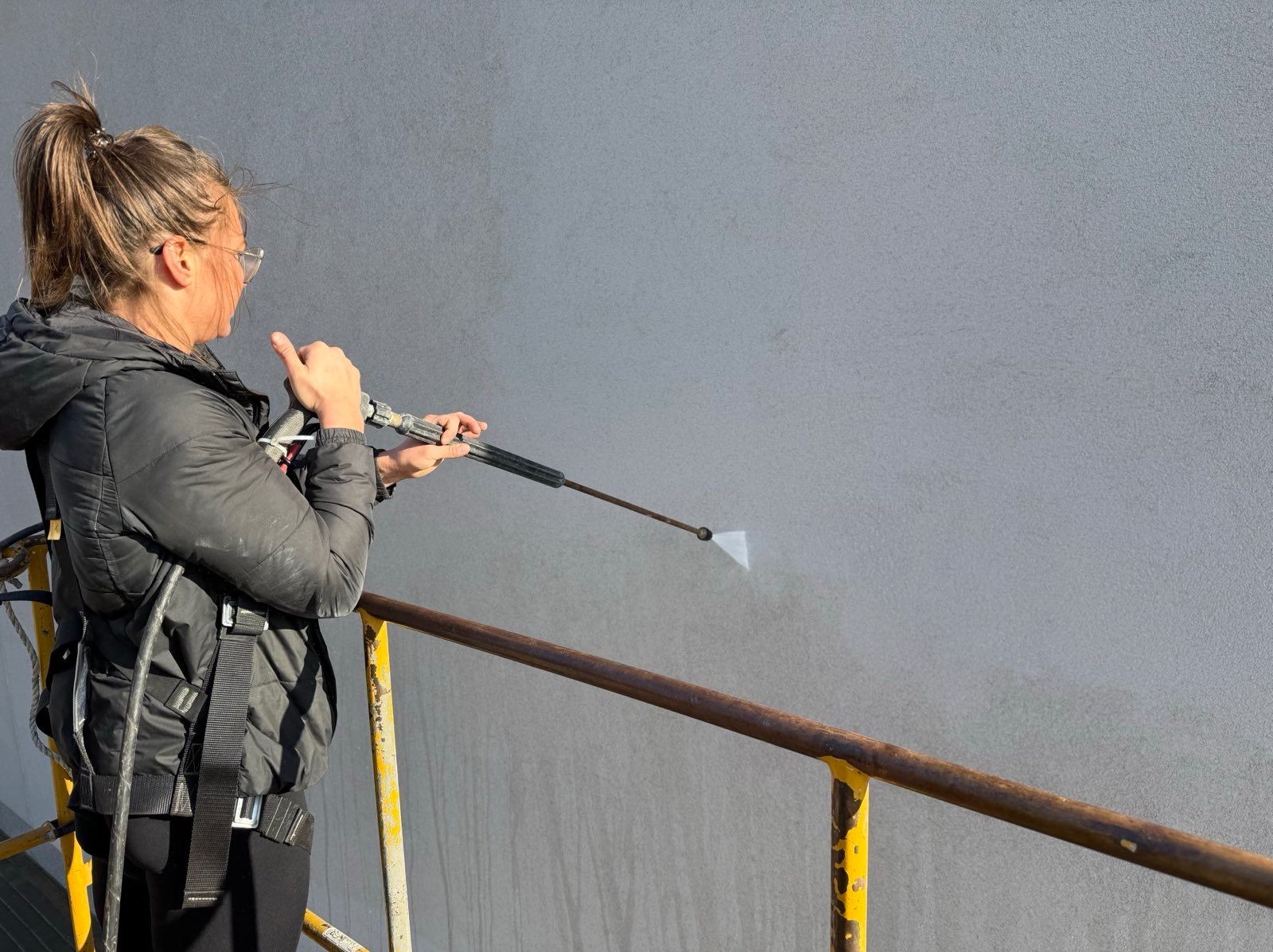 Pracownica w uprzęży, z myjką ciśnieniową, czyści szarą elewację budynku. Widoczne różnice w czystości ściany. Żółta barierka w dolnej części kadru.