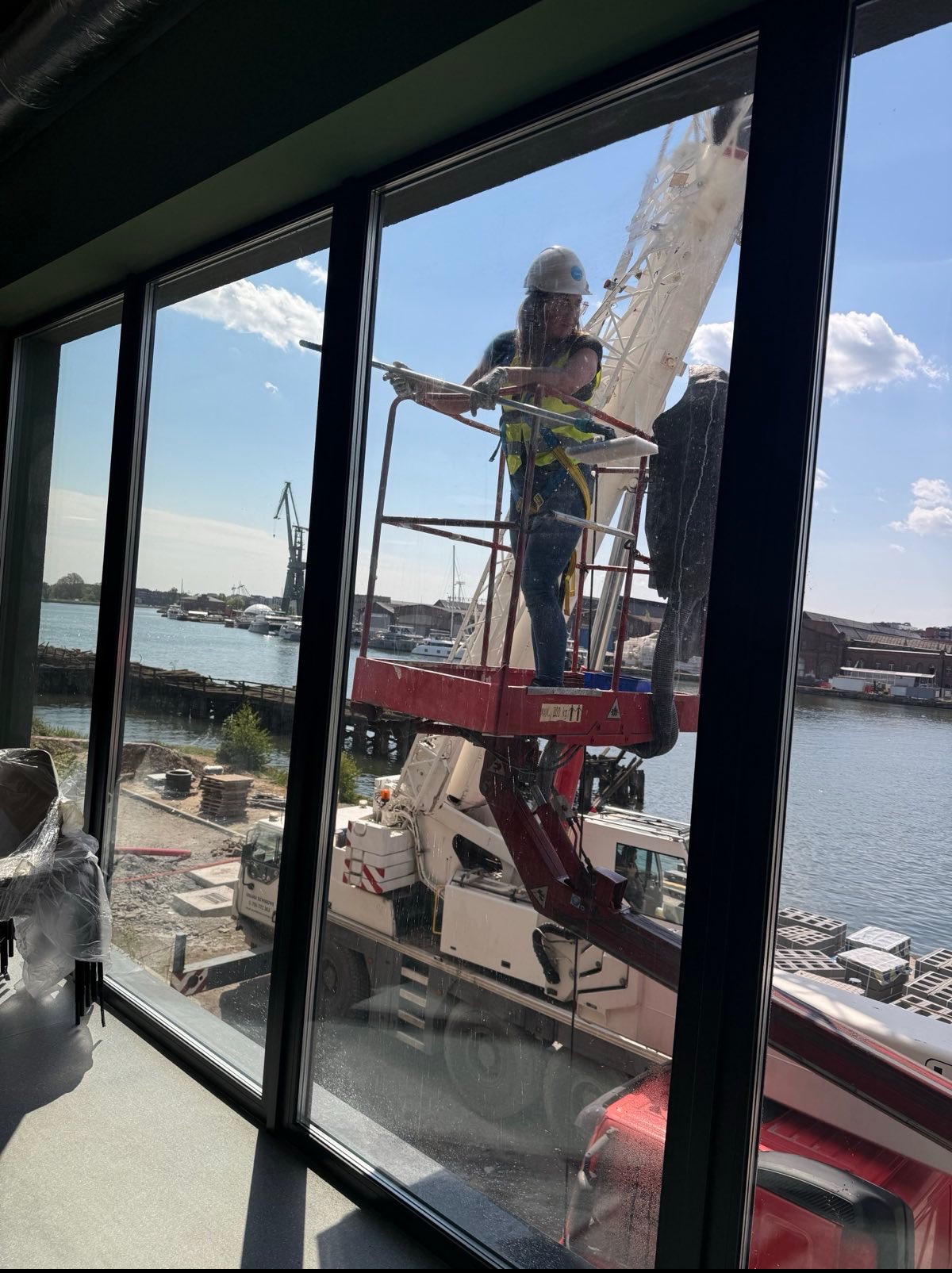 Pracownik w kasku i kamizelce, na podnośniku, myje okna budynku z widokiem na port. W tle widać dźwig portowy i wodę.