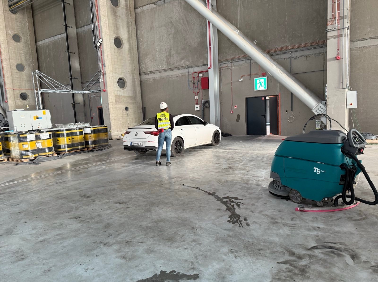 Kobieta w kamizelce nadzoru stoi na posadzce hali obok białego auta i maszyny czyszczącej. W tle widoczne beczki z olejem i betonowe ściany z otworami.