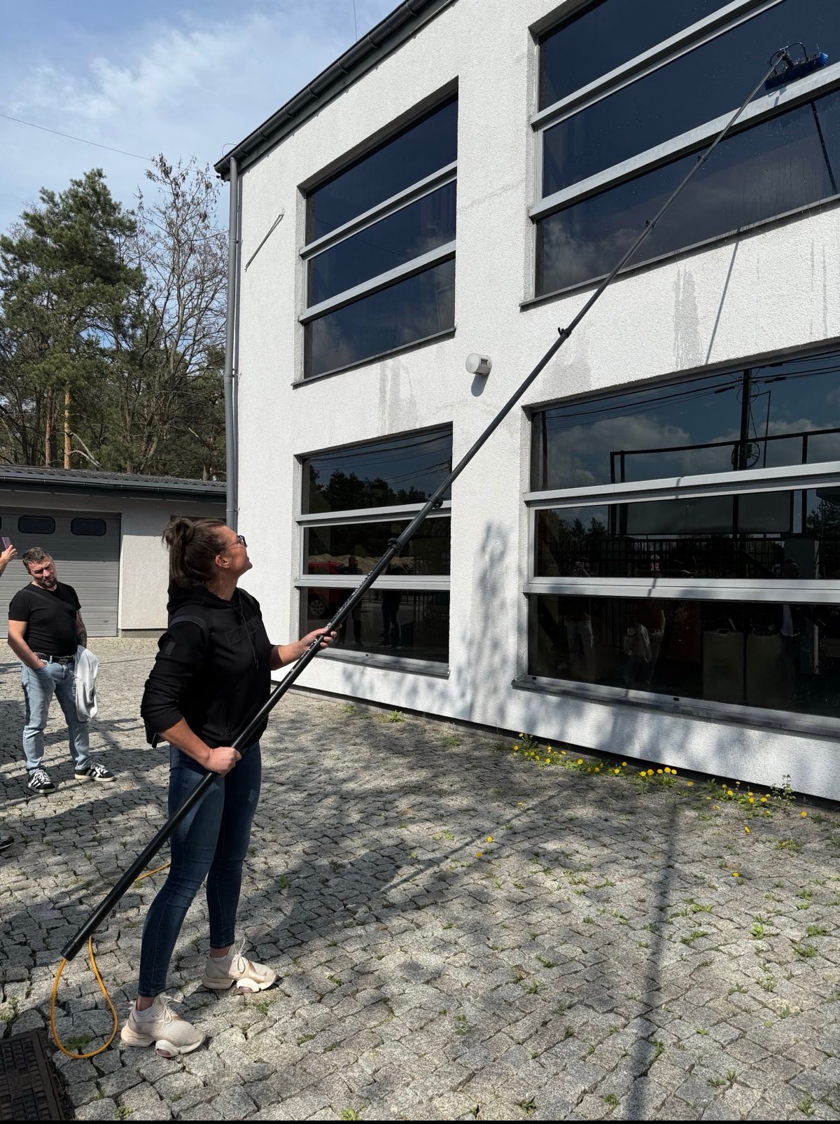 Kobieta myje okna na wysokości za pomocą długiej lancy. Budynek z dużymi oknami, kostka brukowa z żółtymi kwiatami. W tle drzewa i mężczyzna.