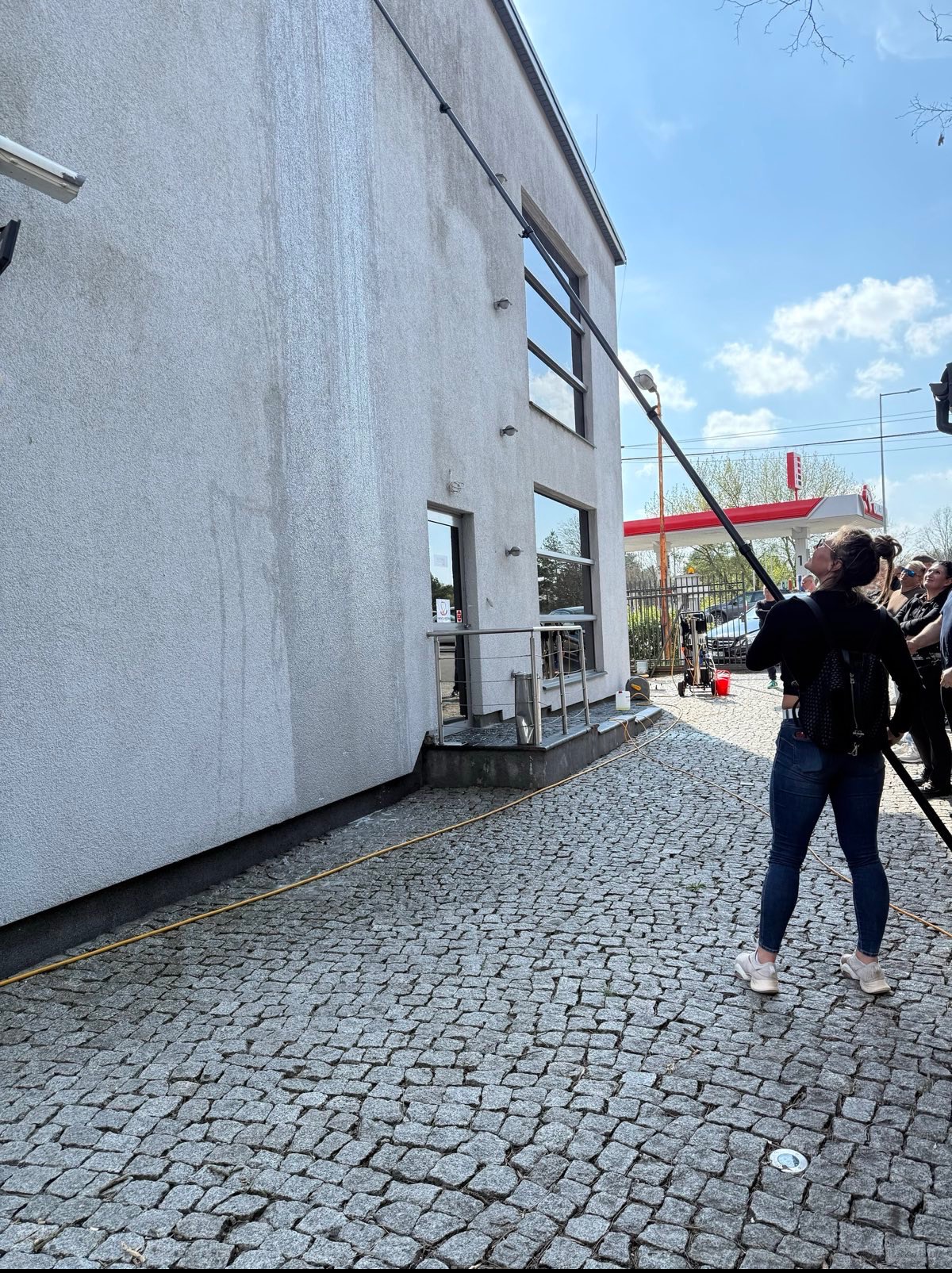 Czyszczenie elewacji budynku z użyciem teleskopowej lancy. Widoczne ślady po czyszczeniu, grupa obserwatorów i oddalona stacja benzynowa w tle.