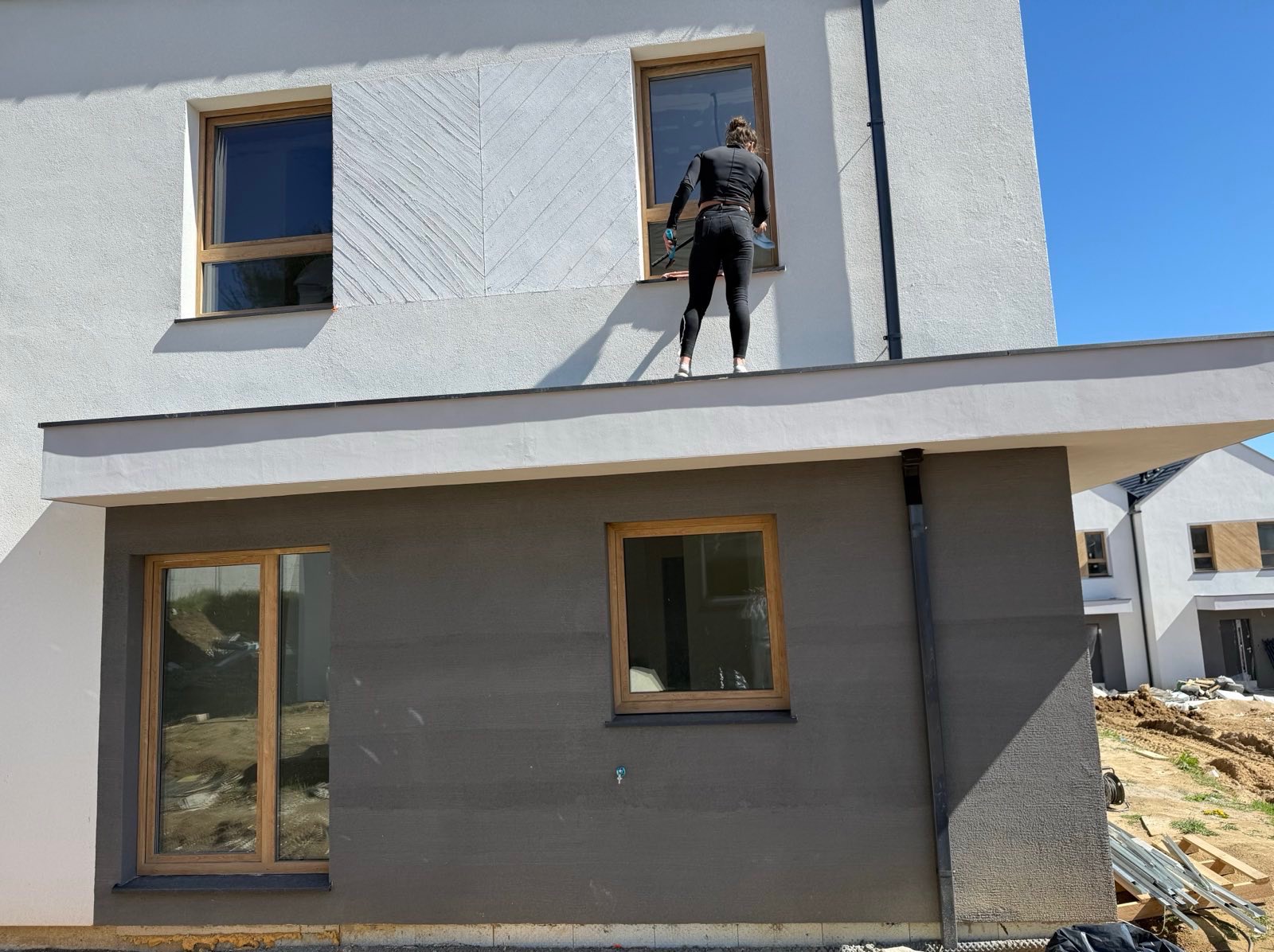 Kobieta w czarnym kombinezonie myje okno na wysokości na tle białego domu z drewnianymi ramami okiennymi i błękitnego nieba.