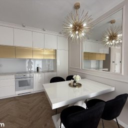 REMAT - Elegancka kuchnia po remoncie w Oleśnicy: białe szafki z złotymi akcentami, parkiet w jodełkę i designerska lampa. Lustro powiększa przestrzeń.