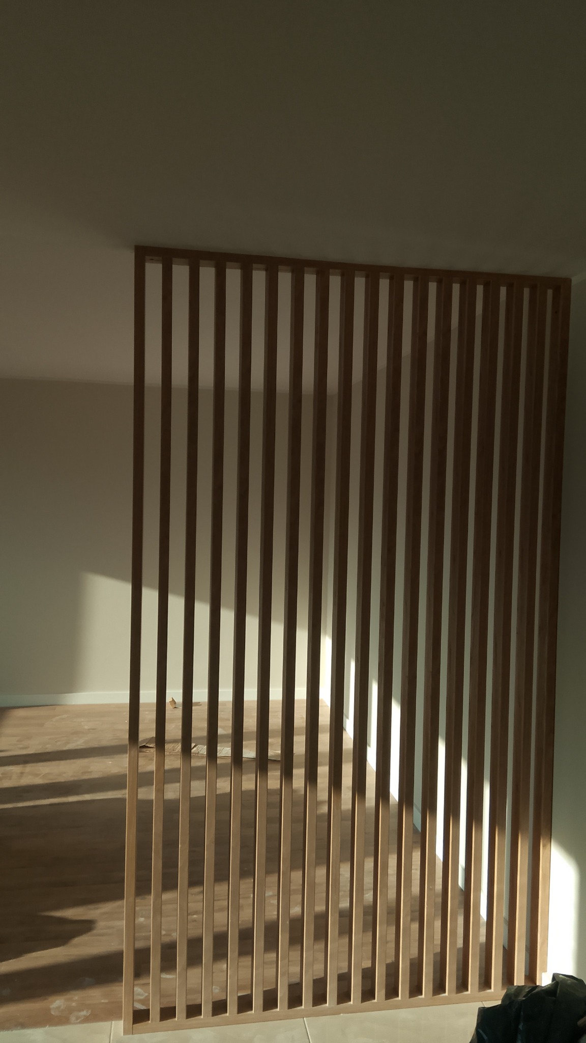 Drewniana ażurowa ścianka działowa w jasnym wnętrzu, z widocznym parkietem i wpadającym światłem słonecznym. Minimalistyczny design, naturalne materiały.