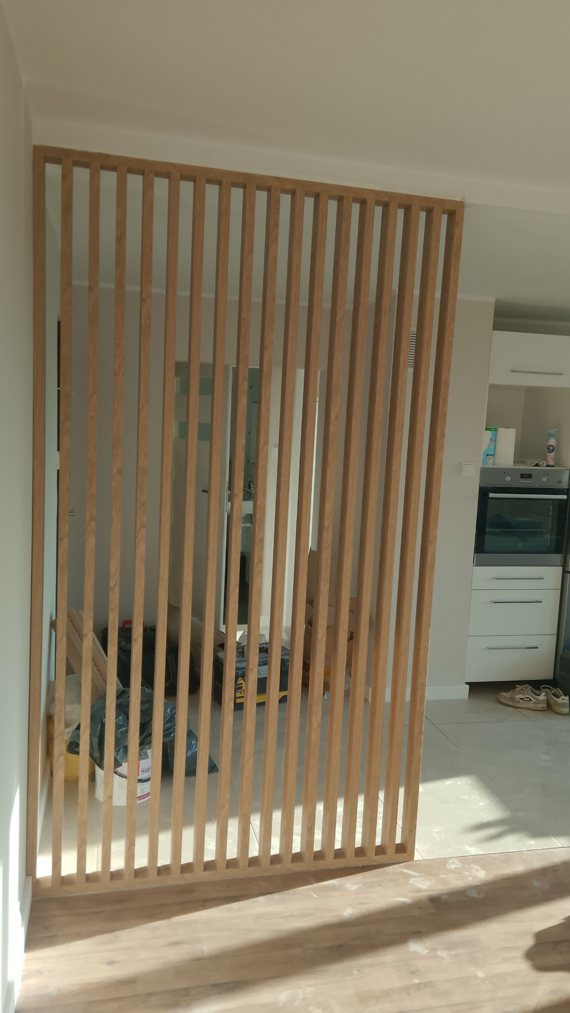 Drewniana ażurowa ścianka działowa z pionowych listew, oddzielająca przestrzeń w jasnym wnętrzu. Widoczny fragment podłogi i elementy wyposażenia kuchni w tle.