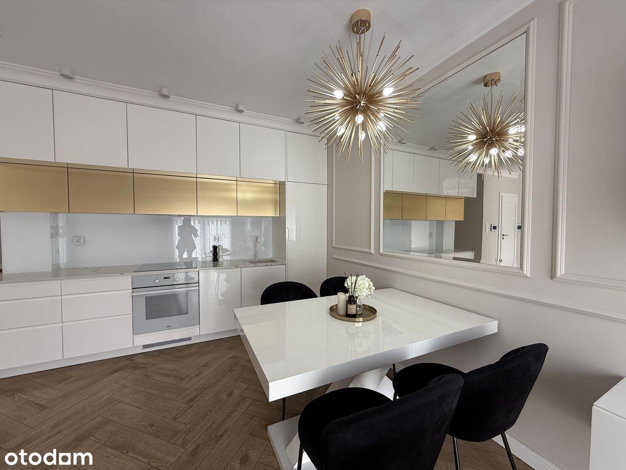 Elegancka kuchnia po remoncie w Oleśnicy: białe szafki z złotymi akcentami, parkiet w jodełkę i designerska lampa. Lustro powiększa przestrzeń.