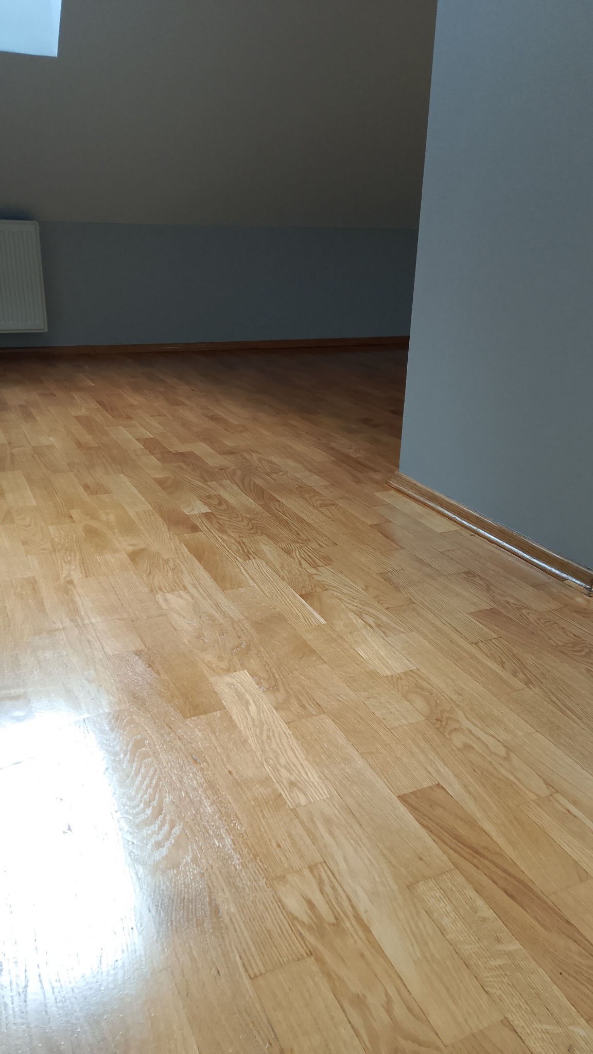 Połyskująca, drewniana podłoga z parkietu w pokoju na poddaszu, świeżo po cyklinowaniu. Szare ściany i biały grzejnik. Ukośny sufit z oknem dachowym.
