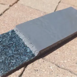 Przekrój warstwowy nowoczesnej dachówki z granulatami, ukazujący jej budowę i strukturę na tle kostki brukowej. Prezentacja materiałów i technologii.