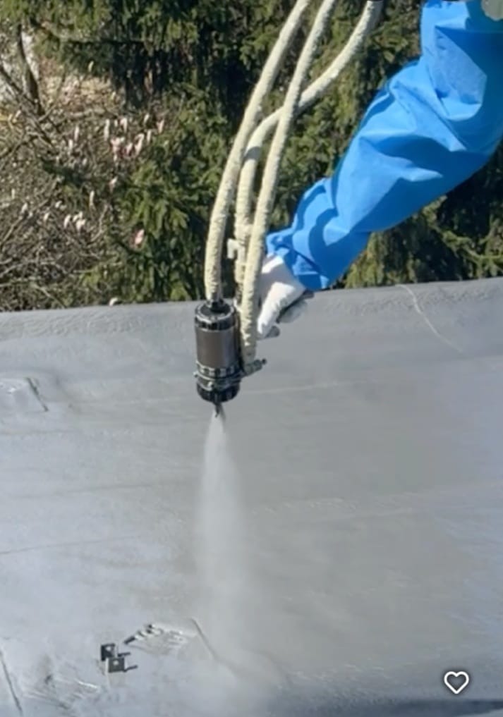 Natrysk izolacji piankowej na płaski dach przez pracownika w niebieskim kombinezonie. Widoczny sprzęt natryskowy i świeżo nałożona warstwa izolacji.