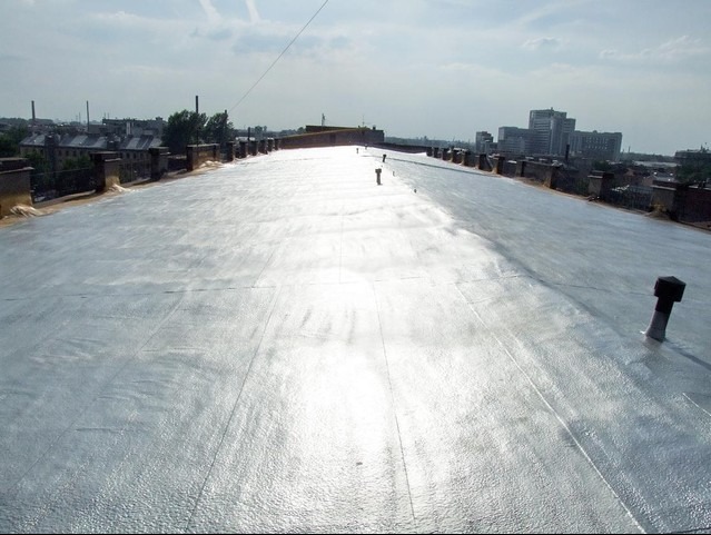 Błyszcząca, srebrzysta izolacja na płaskim dachu budynku miejskiego, widok z góry z kominami i elementami wentylacji. W tle zabudowa miejska i błękitne niebo.