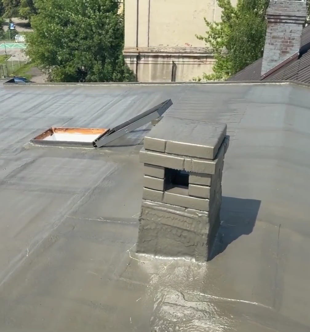 Szary dach po renowacji z kominem i otwartym oknem dachowym. Widoczne ślady napraw i uszczelnienia. Tło stanowią drzewa i budynek.