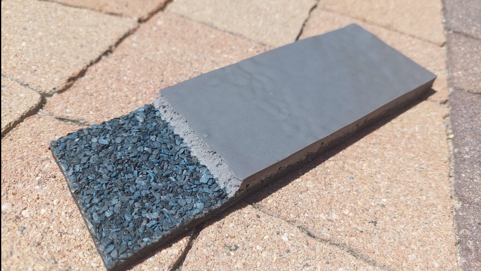 Przekrój warstwowy nowoczesnej dachówki z granulatami, ukazujący jej budowę i strukturę na tle kostki brukowej. Prezentacja materiałów i technologii.