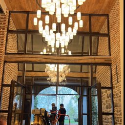 AM ALEKSANDER MIKSZTAL - Montaż designerskiego żyrandola w wysokim holu z ceglanymi ścianami i dużymi oknami. Dwóch monterów na podnośniku. Widok na zewnątrz przez otwarte drzwi.