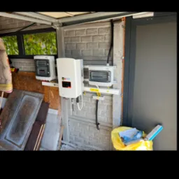 Biały inwerter fotowoltaiczny i dwie skrzynki elektryczne zamontowane na szarej, betonowej ścianie garażu. Widoczne kable i żółte oznaczenia bezpieczeństwa.