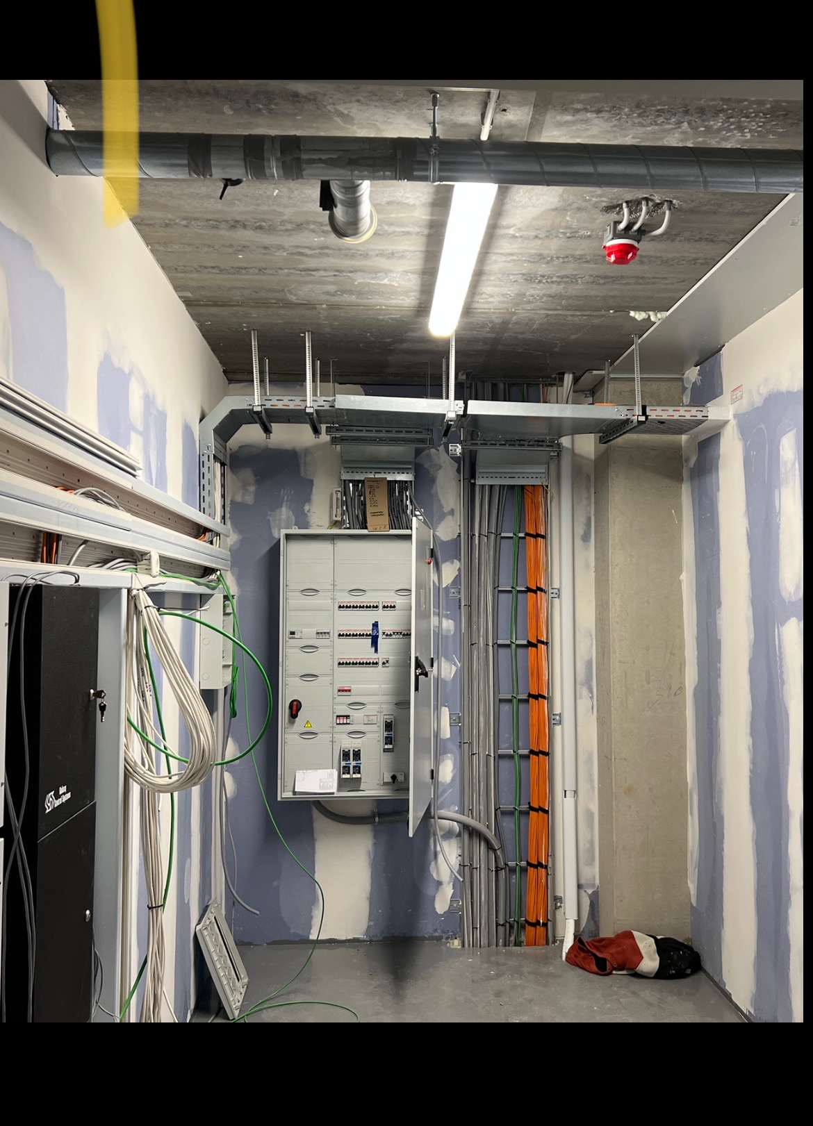 Rozdzielnia elektryczna z otwartymi drzwiami, widoczne kable w korytach i rurach. Ściany w trakcie wykończenia. Oświetlenie sufitowe.