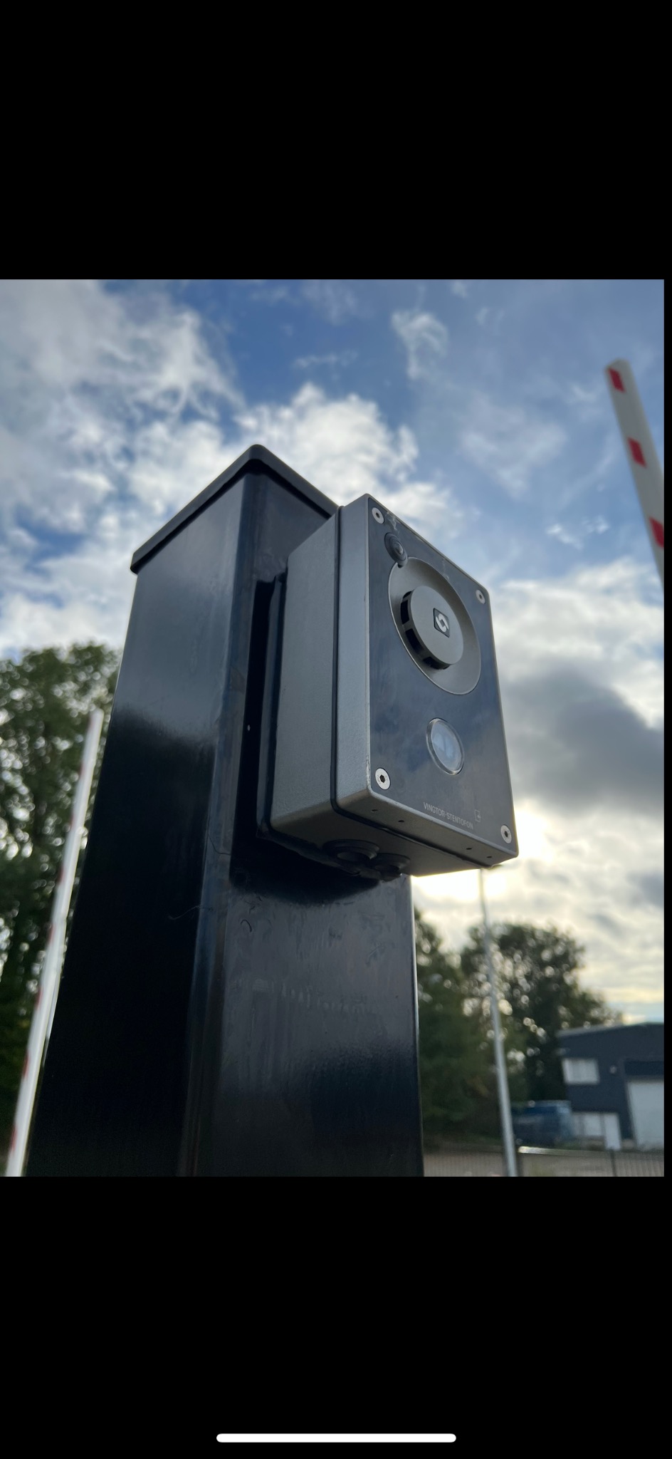Domofon Vingtor-Stentofon zamontowany na słupku przy szlabanie, na tle pochmurnego nieba. Widoczne śruby mocujące i obudowa urządzenia.