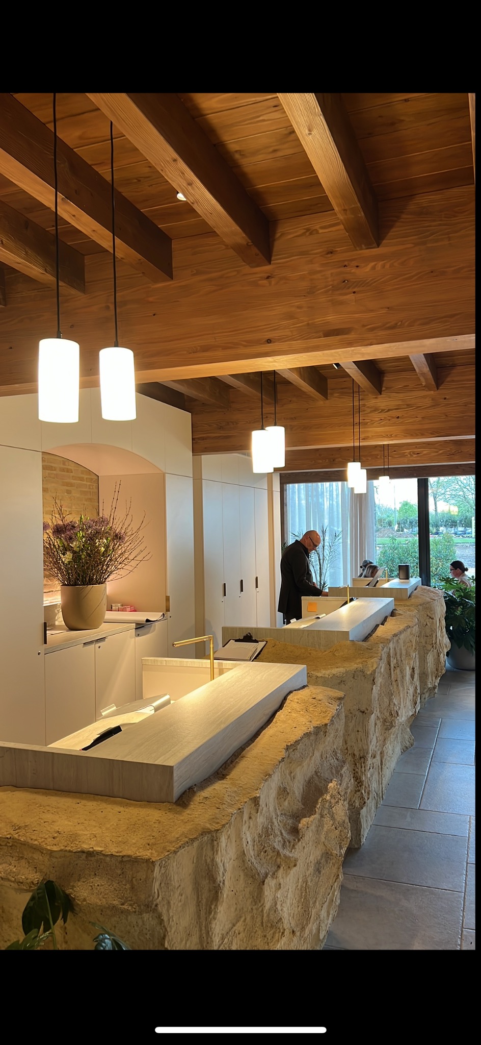 Nowoczesne oświetlenie: cylindryczne lampy wiszące nad kamienną ladą w jasnym wnętrzu z drewnianym sufitem i osobą w tle. Unikalne detale wystroju.