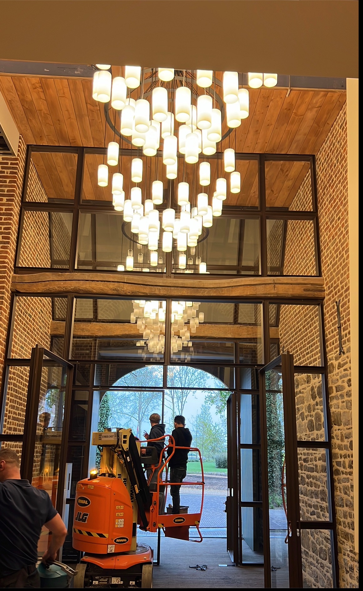Montaż designerskiego żyrandola w wysokim holu z ceglanymi ścianami i dużymi oknami. Dwóch monterów na podnośniku. Widok na zewnątrz przez otwarte drzwi.