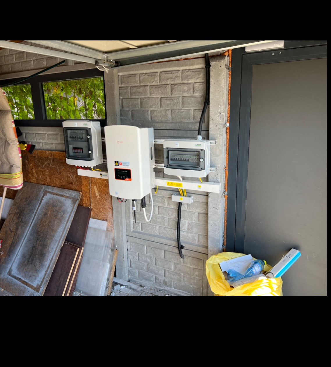 Biały inwerter fotowoltaiczny i dwie skrzynki elektryczne zamontowane na szarej, betonowej ścianie garażu. Widoczne kable i żółte oznaczenia bezpieczeństwa.