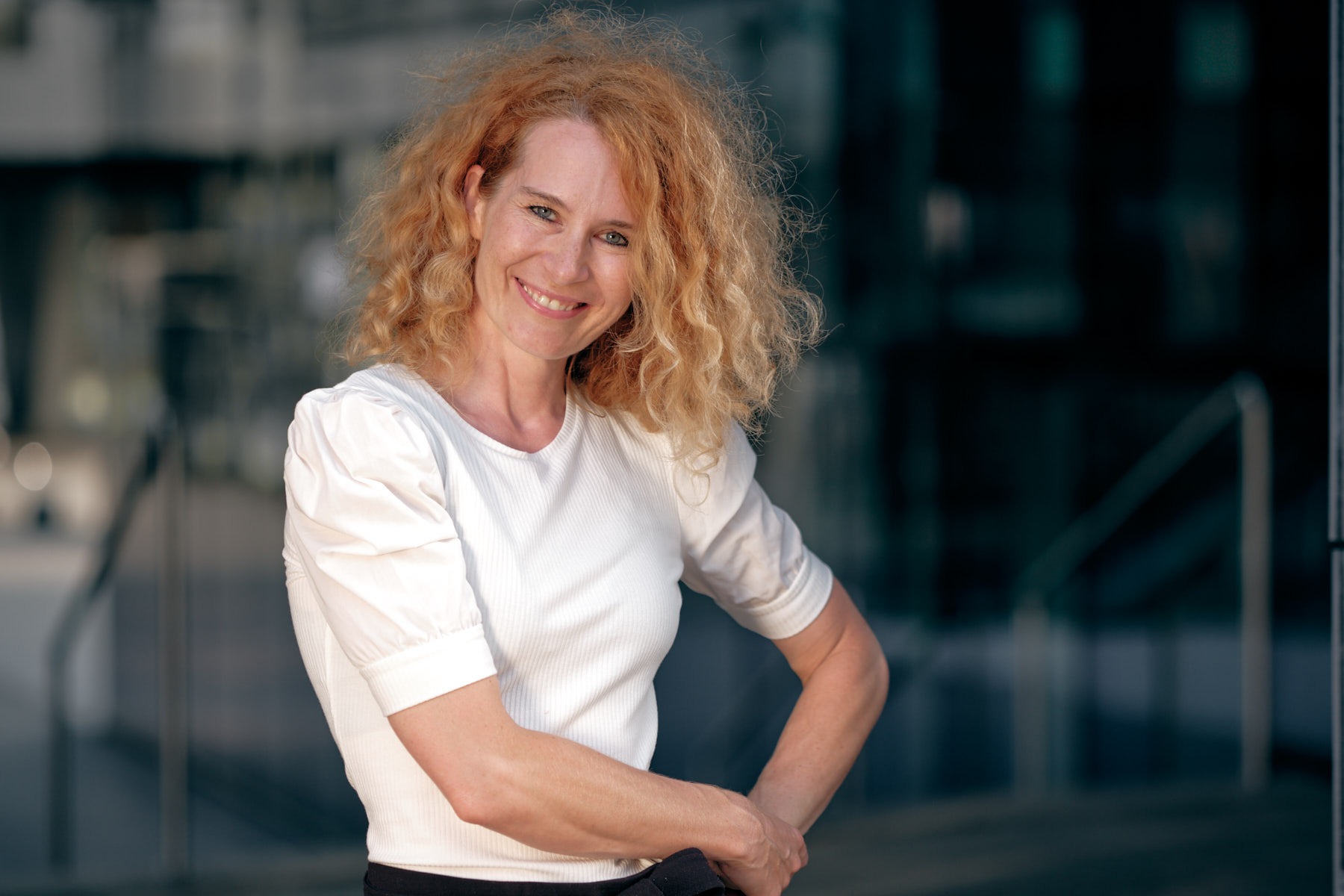 Portret uśmiechniętej kobiety z kręconymi, rudymi włosami w białej bluzce, pozującej na tle nowoczesnego budynku z szklaną fasadą.
