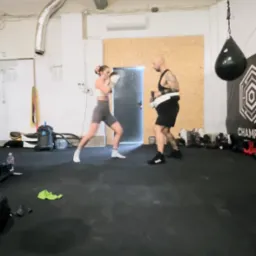 Kobieta w bokserskich rękawicach trenuje z trenerem na sali. W tle worek treningowy i sprzęt. Dynamiczne ujęcie z treningu sportów walki.