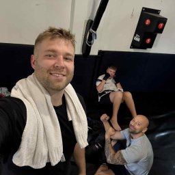 Łukasz Kudła - Trener personalny robi selfie z podopiecznymi w sali treningowej. Widoczny sprzęt do ćwiczeń i mata. Jeden z podopiecznych ćwiczy pod okiem trenera.