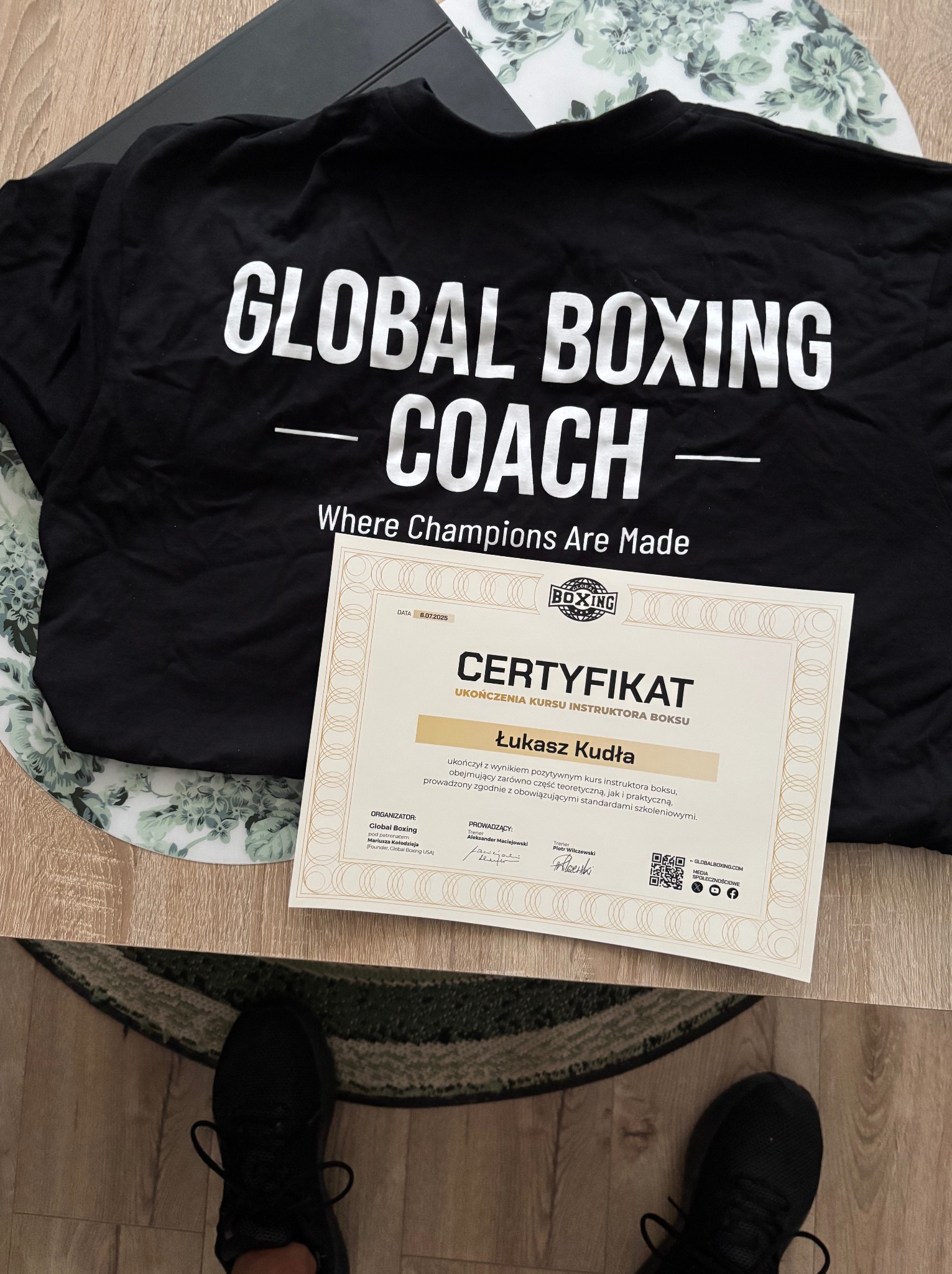 Certyfikat ukończenia kursu instruktora boksu 'Global Boxing Coach' leży na czarnej koszulce z logo, widoczne buty trenera. Data ważności 06.07.2025.