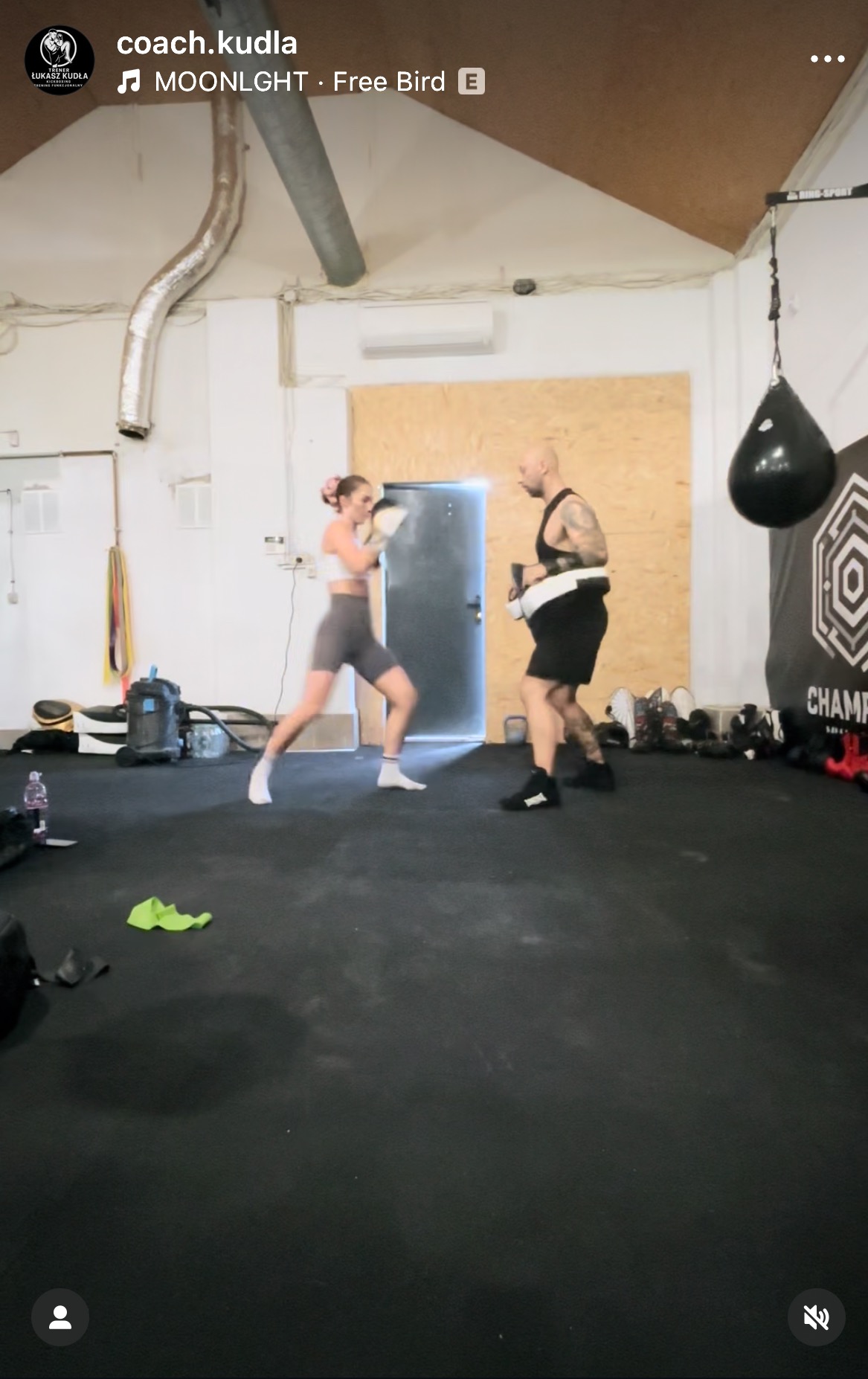 Kobieta w bokserskich rękawicach trenuje z trenerem na sali. W tle worek treningowy i sprzęt. Dynamiczne ujęcie z treningu sportów walki.