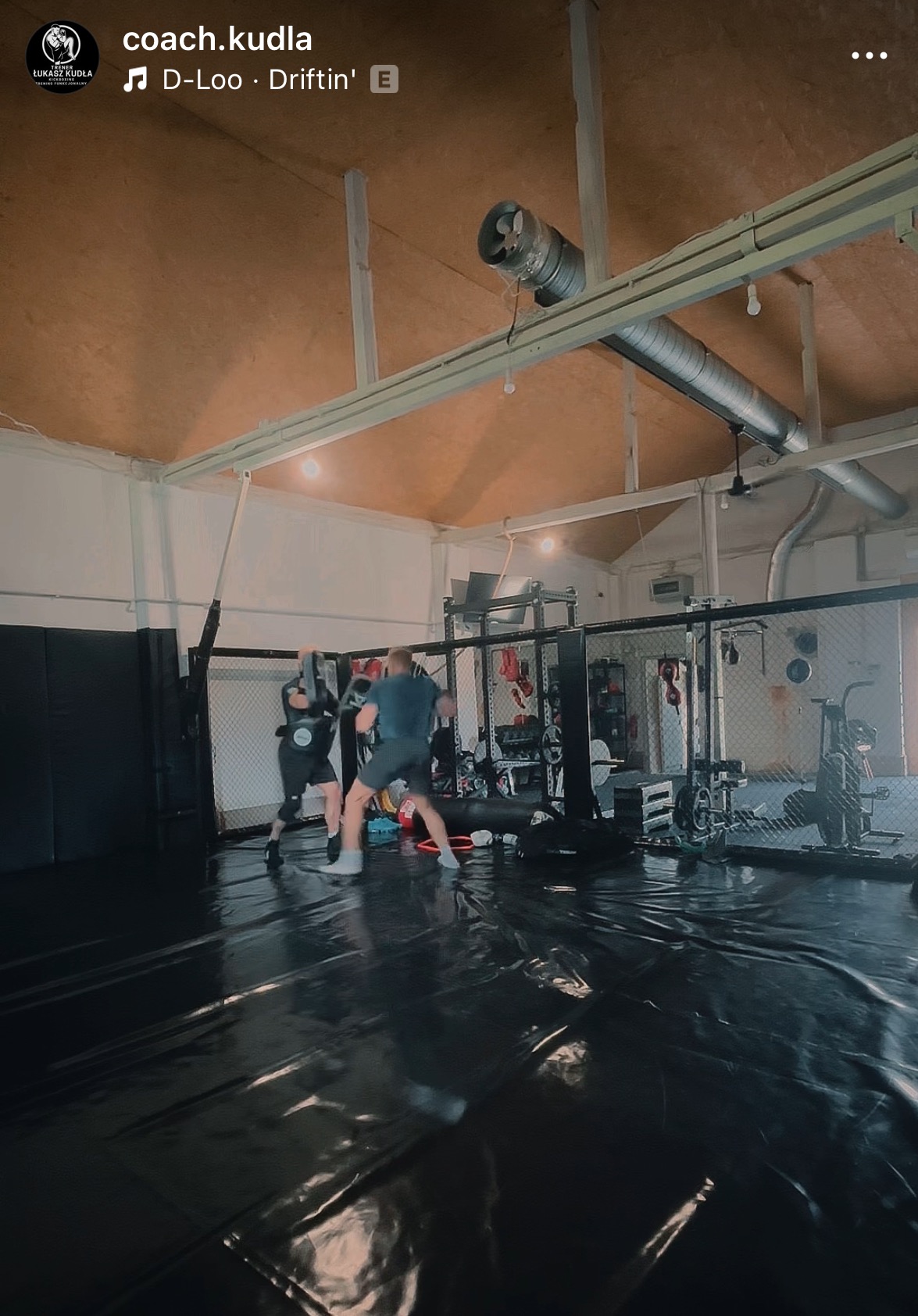 Trening sportów walki: sparing partnerzy w akcji na czarnej macie w sali z klatką MMA i sprzętem do ćwiczeń w tle. Dynamiczne ujęcie, akcent na ruch.