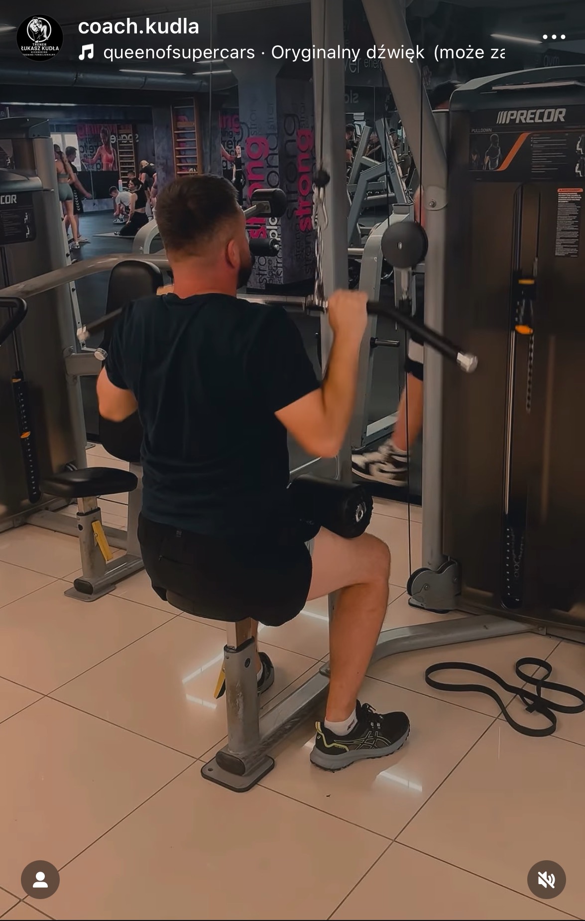 Mężczyzna ćwiczy na maszynie do wyciągania drążka w dół, w tle widoczne inne osoby w siłowni. Skupienie na plecach i ramionach ćwiczącego.