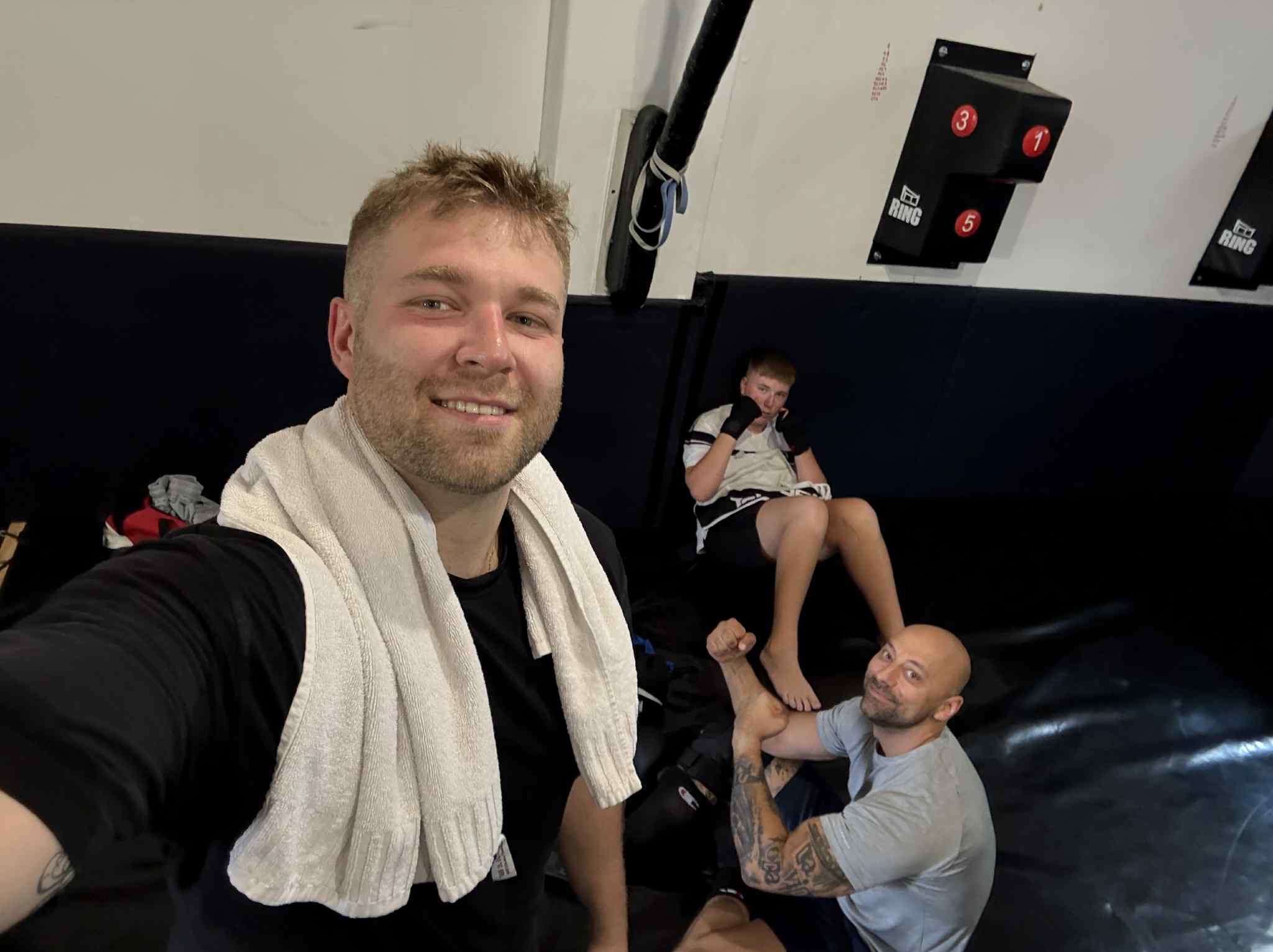 Trener personalny robi selfie z podopiecznymi w sali treningowej. Widoczny sprzęt do ćwiczeń i mata. Jeden z podopiecznych ćwiczy pod okiem trenera.