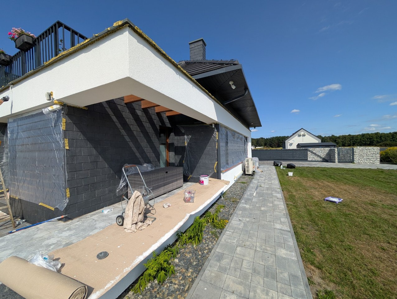 Elewacja domu w trakcie malowania na czarno, zabezpieczona folią. Widoczne narzędzia, trawa i kostka brukowa. Słoneczny dzień, błękitne niebo.