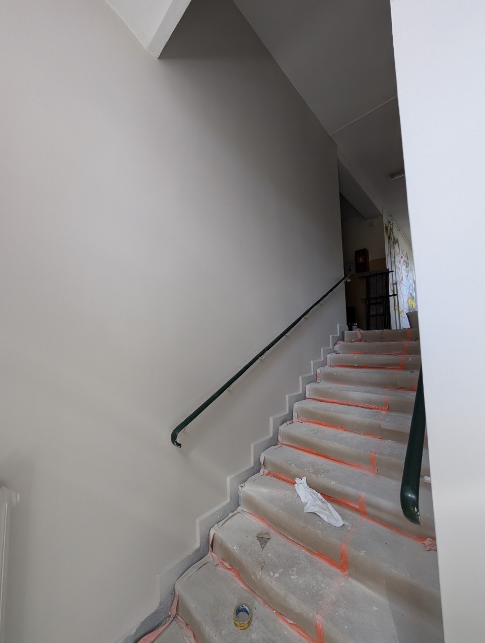 Jasne, świeżo pomalowane ściany na klatce schodowej z zieloną poręczą. Schody zabezpieczone folią malarską, widoczny pędzel i ścierka. Ujęcie z dołu.