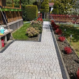 Łukasz Sarba LuClean - Jasna kostka brukowa na ścieżce ogrodowej, obrzeża z ciemnego grysu, rabaty z roślinami. Widok na dom z drewnianą werandą i zadbany ogród.