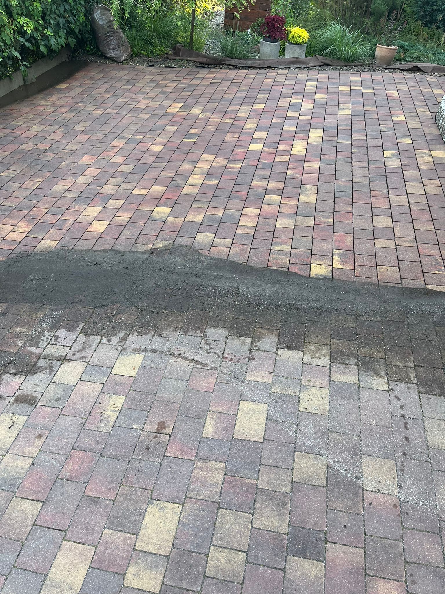 Częściowo umyta kostka brukowa w odcieniach czerwieni i żółci, kontrastująca z brudną powierzchnią. Widoczne rośliny i worki w tle.