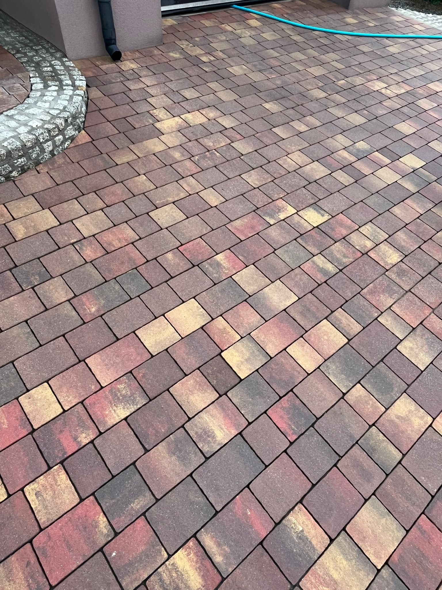 Wielobarwna kostka brukowa ułożona w jodełkę, z elementami obrzeża z kamienia polnego i fragmentem węża ogrodowego w tle.