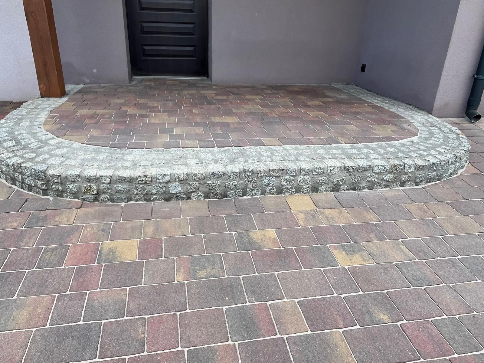Ułożona kostka brukowa w odcieniach brązu i beżu, tworząca ścieżkę przed wejściem do budynku, z obrzeżem z szarej kostki granitowej.