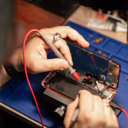 Pośrednictwo Finansowe Igor Kalinowski - Serwisant w trakcie diagnostyki smartfona miernikiem; rozłożony telefon na gumowej macie serwisowej, widoczne narzędzia i akcesoria; ujęcie z góry.