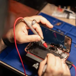 Serwisant w trakcie diagnostyki smartfona miernikiem; rozłożony telefon na gumowej macie serwisowej, widoczne narzędzia i akcesoria; ujęcie z góry.