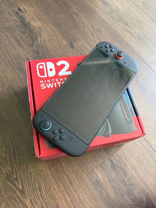 Czarna konsola do gier na czerwonym pudełku z logo na drewnianej podłodze, leżąca pod kątem.