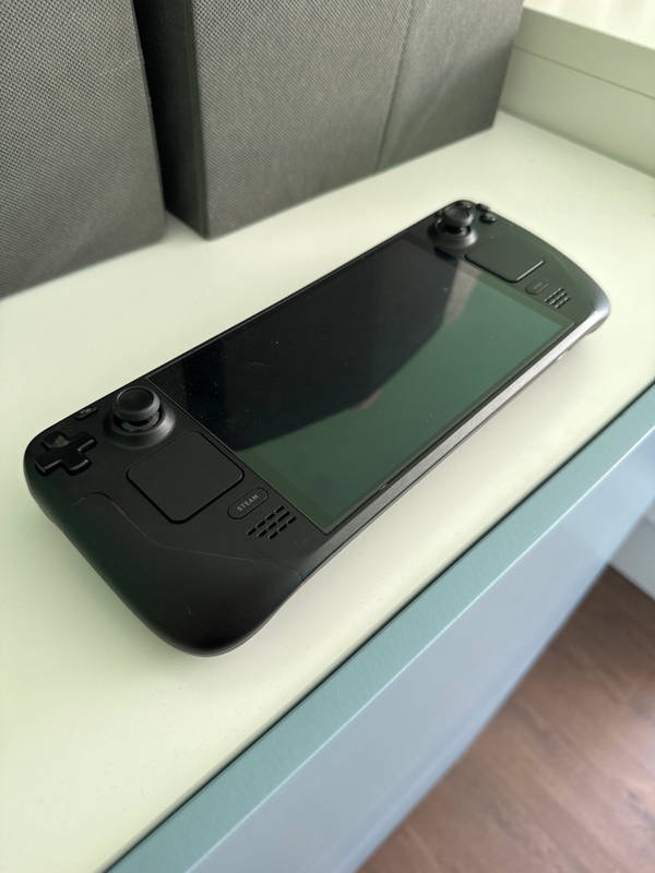 Czarna konsola przenośna Steam Deck leży na białym blacie, z szarymi pudełkami w tle. Widoczne analogi, przyciski i ekran. Całość w neutralnym oświetleniu.