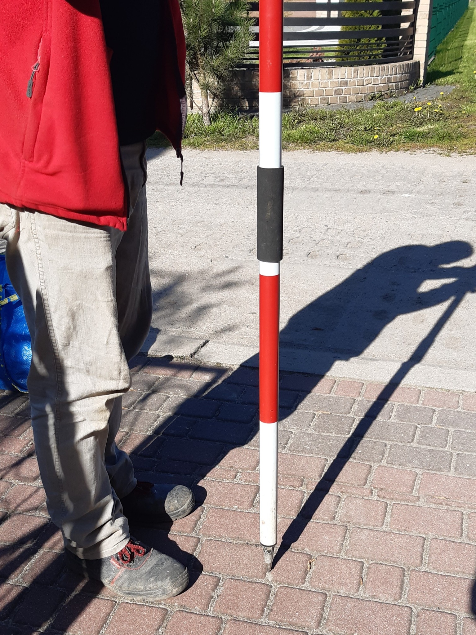 Geodeta w czerwonej bluzie, trzymający łatę geodezyjną na chodniku z kostki brukowej, cień osoby i narzędzia na chodniku, w tle ogrodzenie z cegły.
