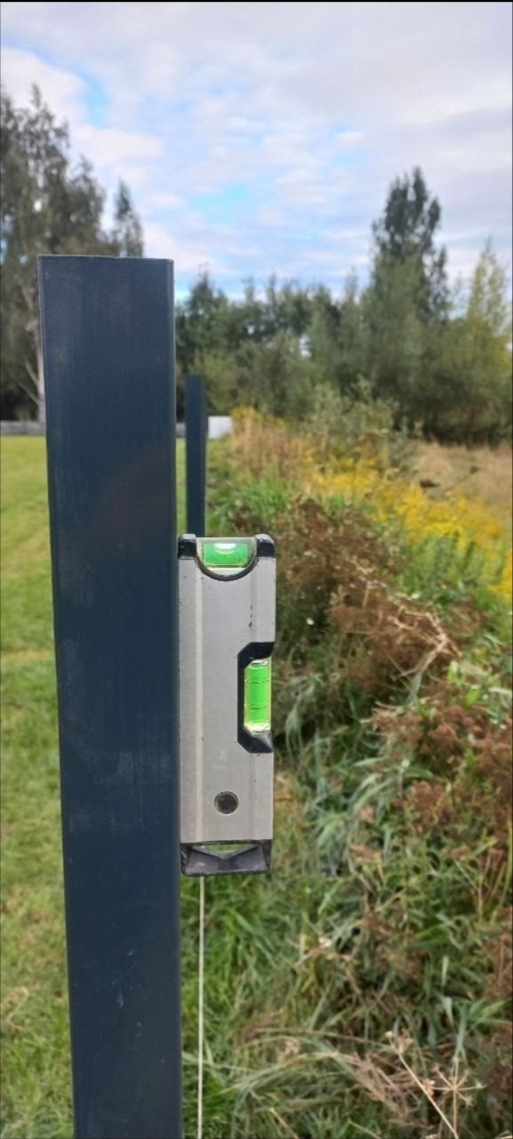 Poziomnica na słupku ogrodzenia panelowego, w tle łąka i drzewa. Detal montażu ogrodzenia panelowego, weryfikacja pionu słupka.