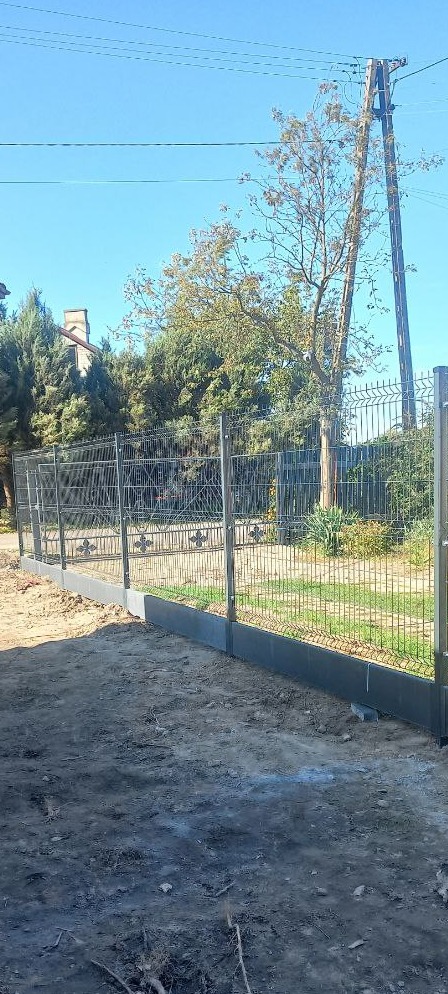 Szare ogrodzenie panelowe z podmurówką na nierównym terenie, widoczne słupy energetyczne w tle, słoneczny dzień.