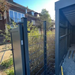 Nowoczesna, grafitowa brama panelowa z furtką, widoczna na tle domu z cegły i zieleni. W tle wiata śmietnikowa z poziomymi panelami.