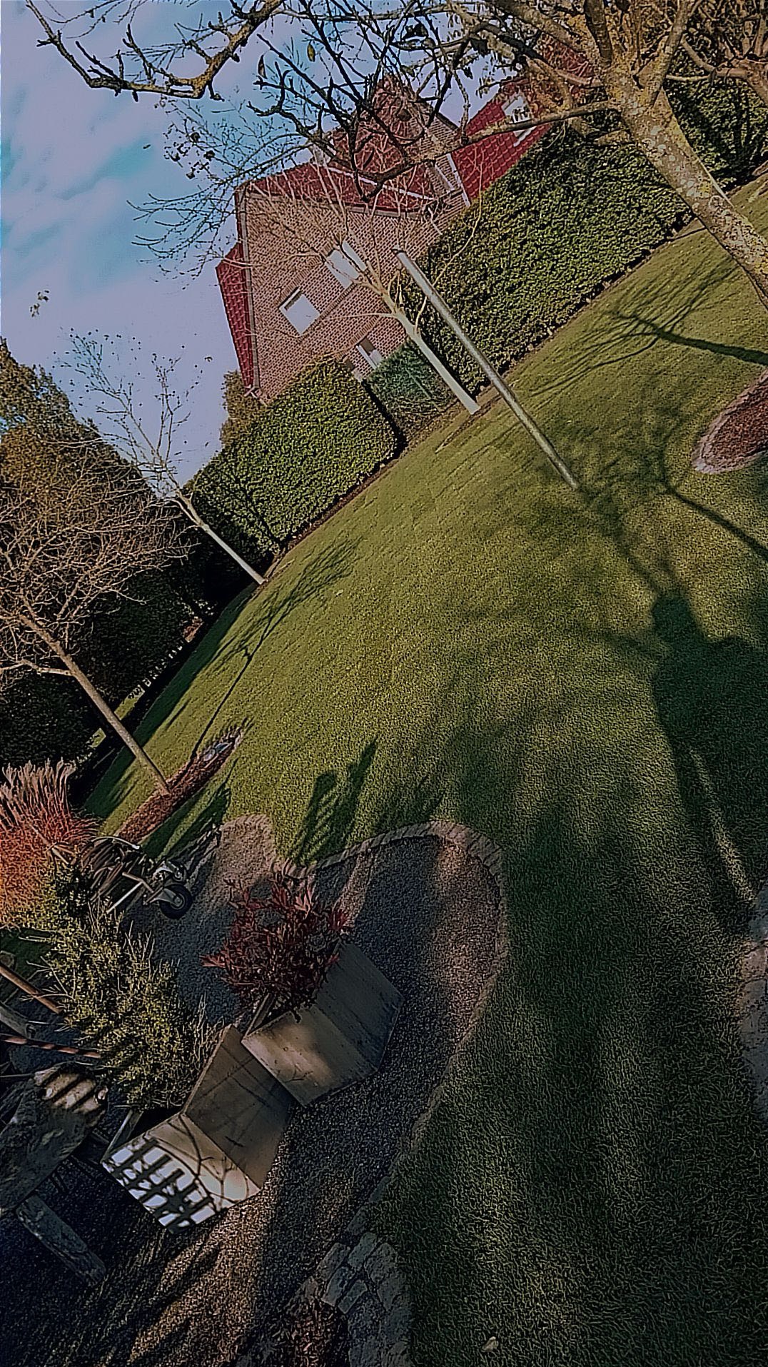 Zadbany trawnik z równo przyciętą trawą i żywopłotem, obok domu z czerwonej cegły. Widoczne drzewa i cień osoby. Kąt ujęcia z góry podkreśla geometrię ogrodu.