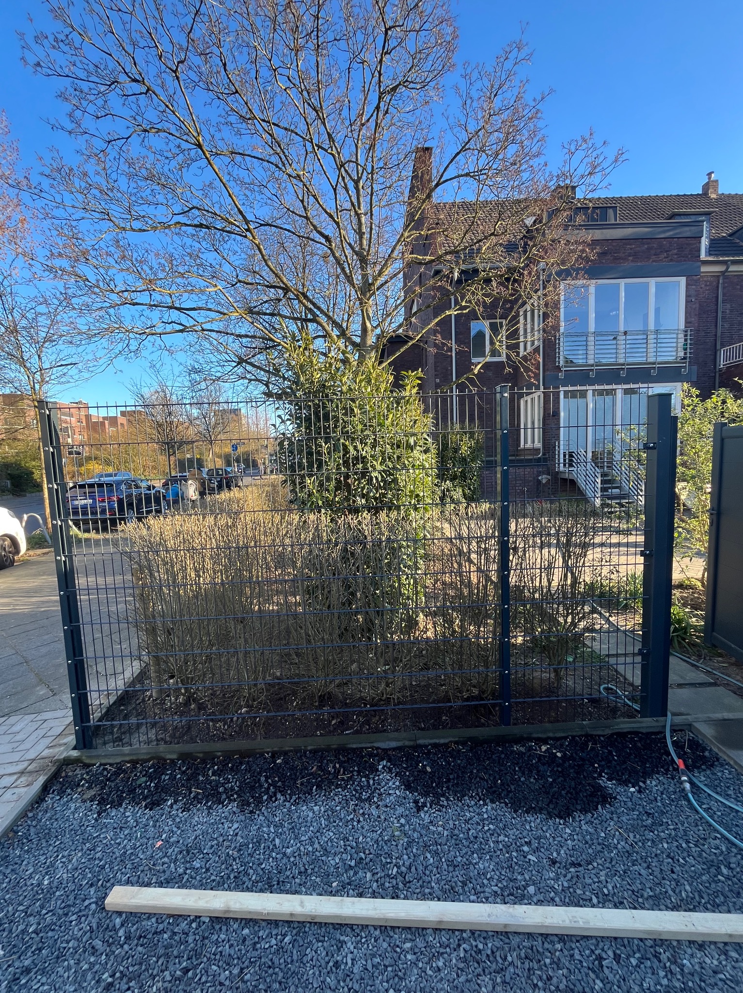 Nowoczesne ogrodzenie panelowe z metalu, oddzielające posesję od ulicy. Widoczne krzewy, drzewo i fragment elewacji budynku w tle. Żwirowe podłoże.