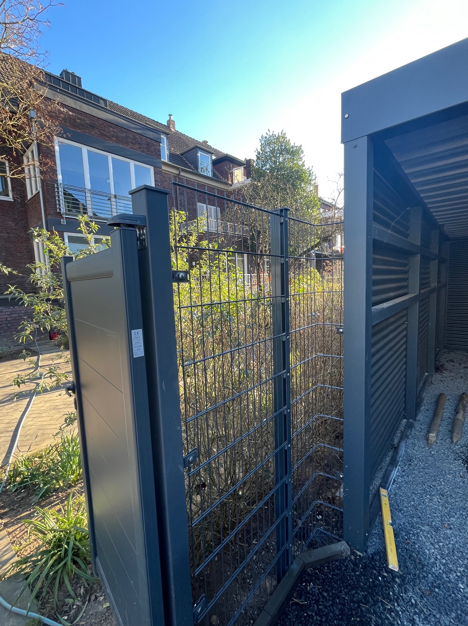 Nowoczesna, grafitowa brama panelowa z furtką, widoczna na tle domu z cegły i zieleni. W tle wiata śmietnikowa z poziomymi panelami.