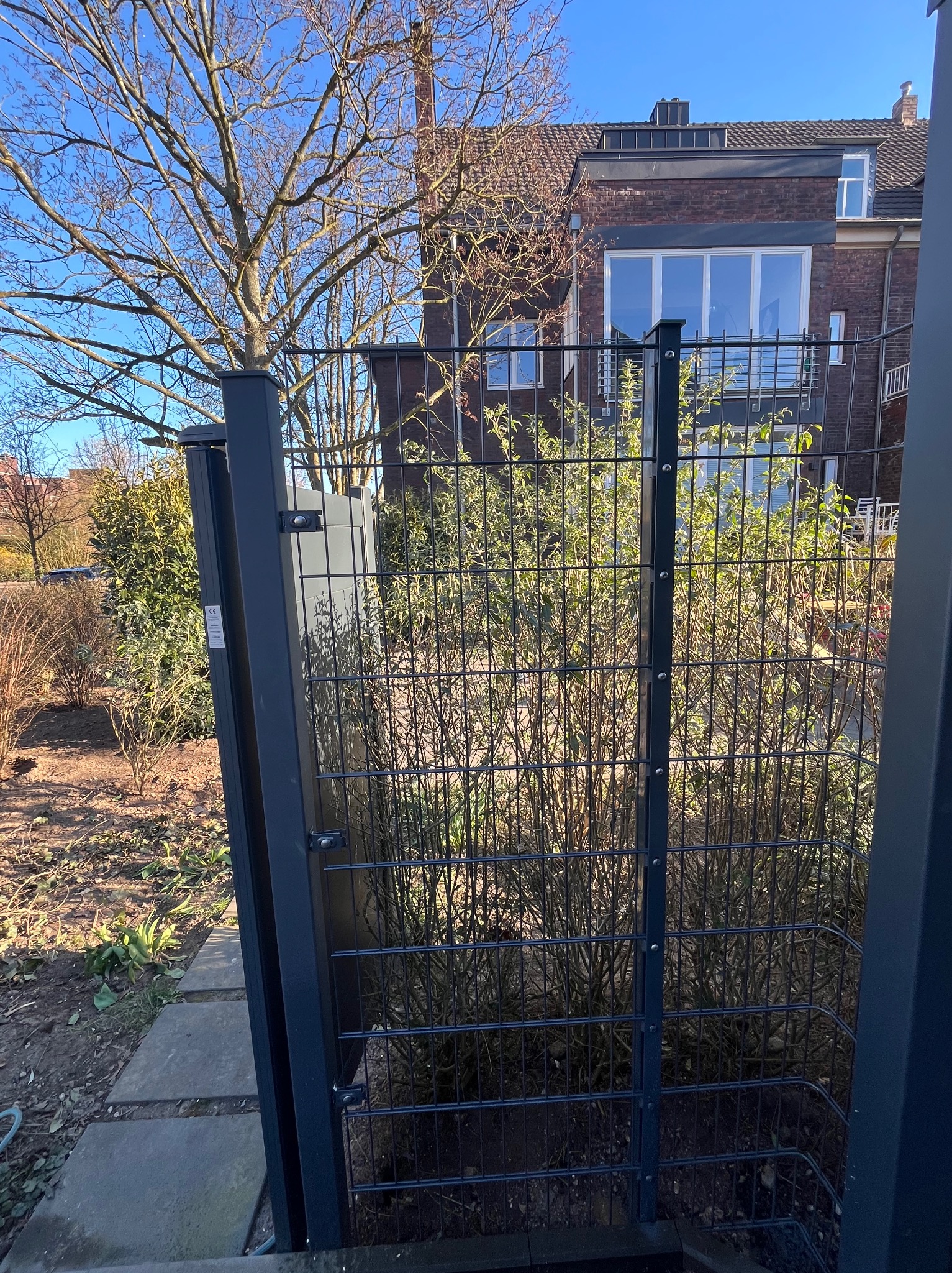 Nowoczesne ogrodzenie panelowe z bramką, widok z bliska na tle domu z cegły i ogrodu z drzewami. Metalowa konstrukcja w kolorze grafitowym.