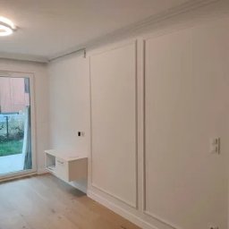 Studio52, Kamil Mistrzak - Jasne wnętrze po remoncie: białe ściany z dekoracyjnymi panelami, drewniana podłoga, przesuwne drzwi balkonowe i minimalistyczna szafka wisząca. Oświetlenie sufitowe.